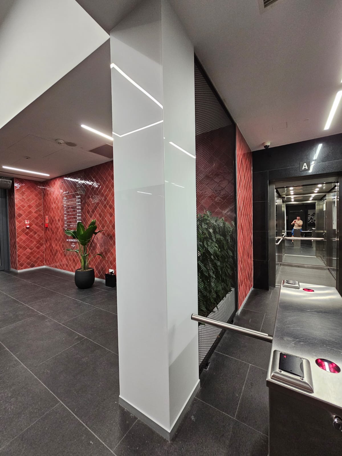 Nowoczesne wnętrze biurowca z przeszkloną kolumną i elementami dekoracyjnymi. Widoczny fragment windy i roślinność w tle. Oświetlenie LED podkreśla detale.