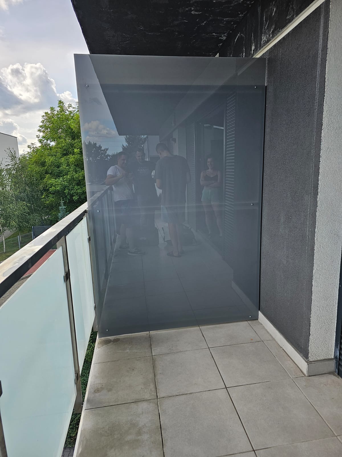 Szklana zabudowa balkonu z widocznymi mocowaniami, odbijająca sylwetki osób wewnątrz. Nowoczesne rozwiązanie architektoniczne z szarymi płytkami podłogowymi.
