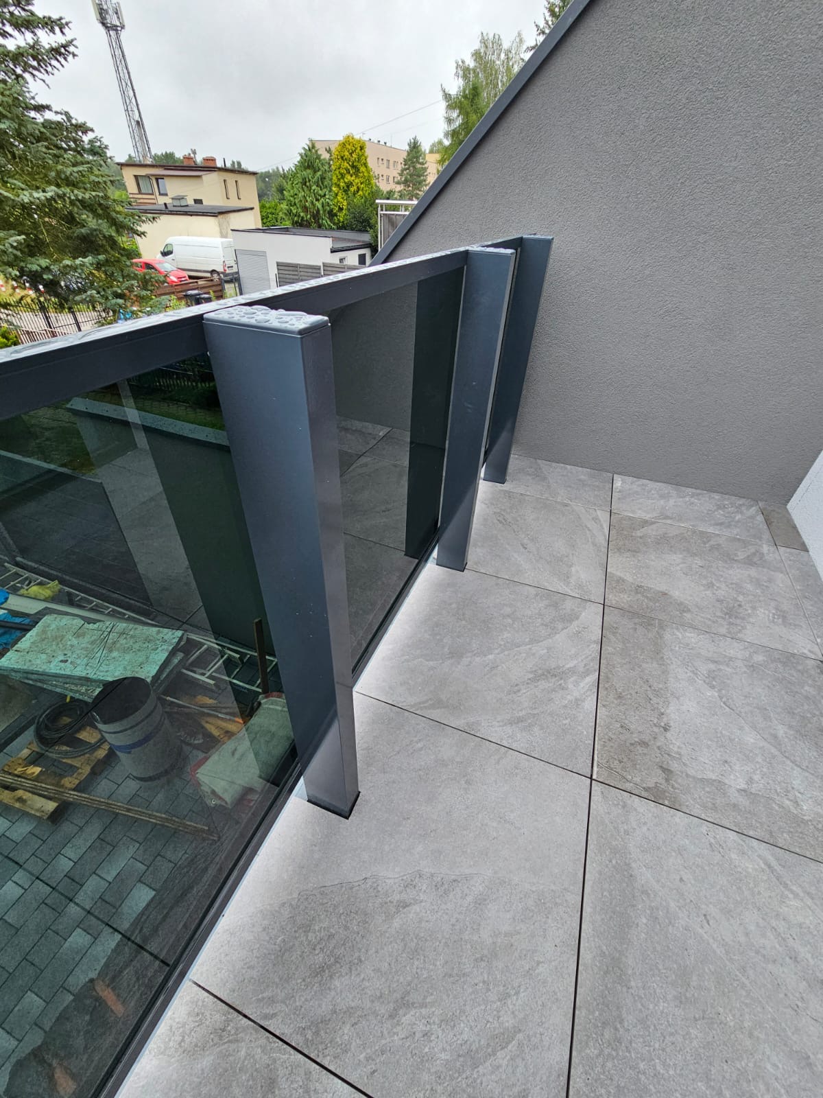 Szklana balustrada balkonowa z grafitowymi słupkami, widok z góry na szare płytki podłogowe i otoczenie budynku w tle. Nowoczesny design.
