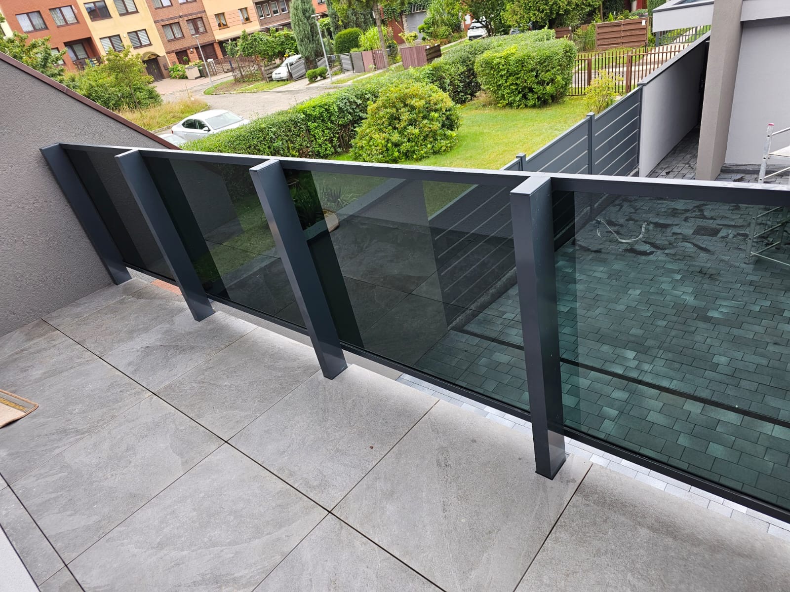 Nowoczesna balustrada ze szkła grafitowego z ciemną ramą na balkonie z szarymi płytkami. Widok na zadbany ogród i budynki mieszkalne w tle.