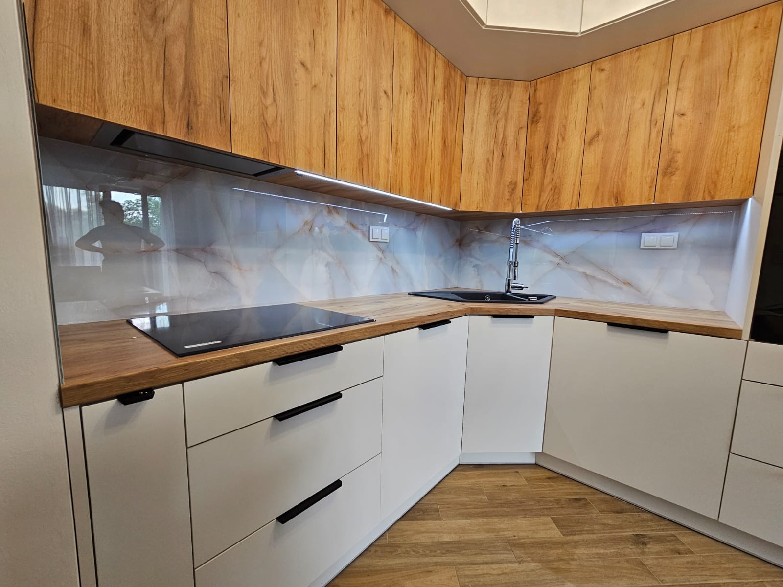 Nowoczesna kuchnia z drewnianymi szafkami górnymi i białymi dolnymi. Marmurowy panel nad blatem. Widoczne odbicie osoby w szklanym panelu.