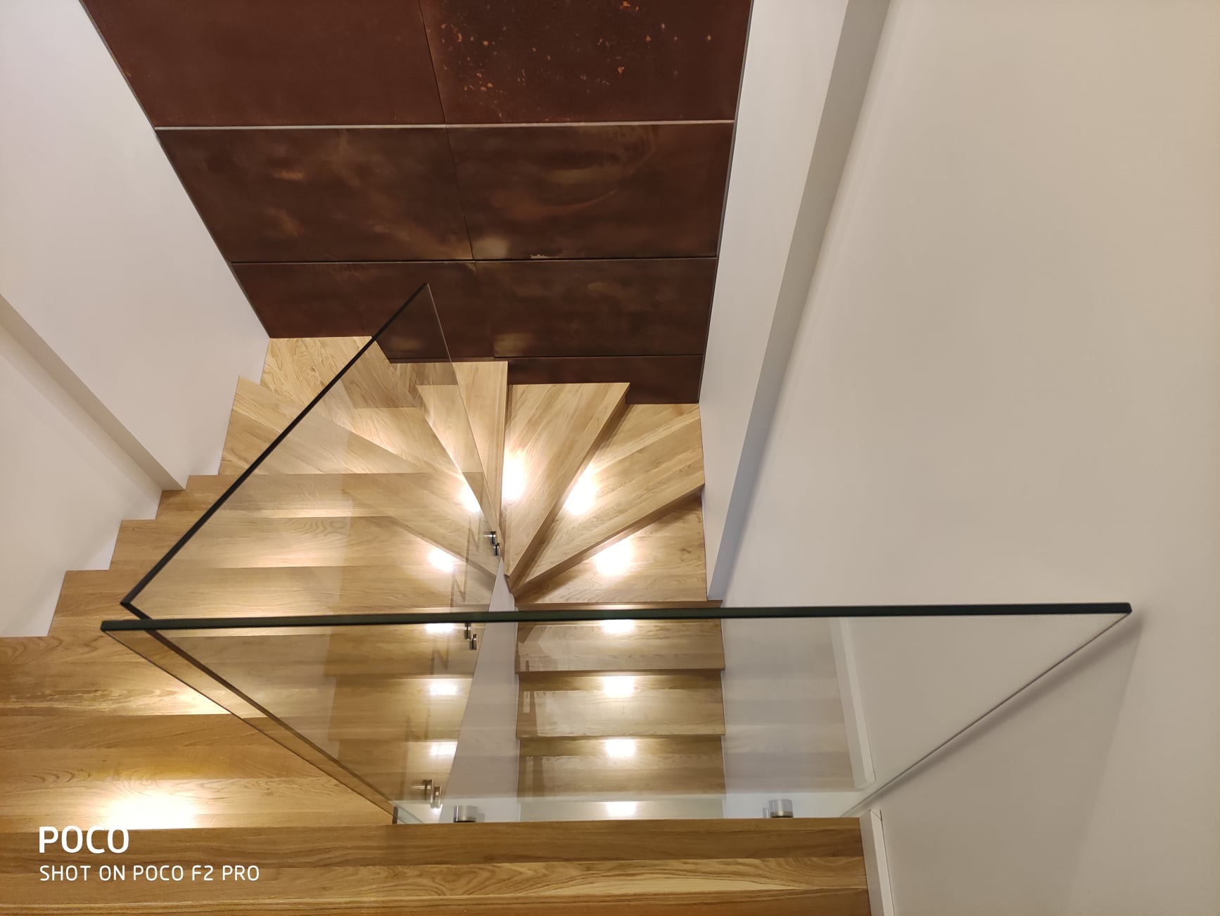 Spiralne schody z jasnego drewna zabezpieczone nowoczesną, szklaną balustradą z czarnymi elementami wykończeniowymi, widziane z góry, z brązowym sufitem w tle.