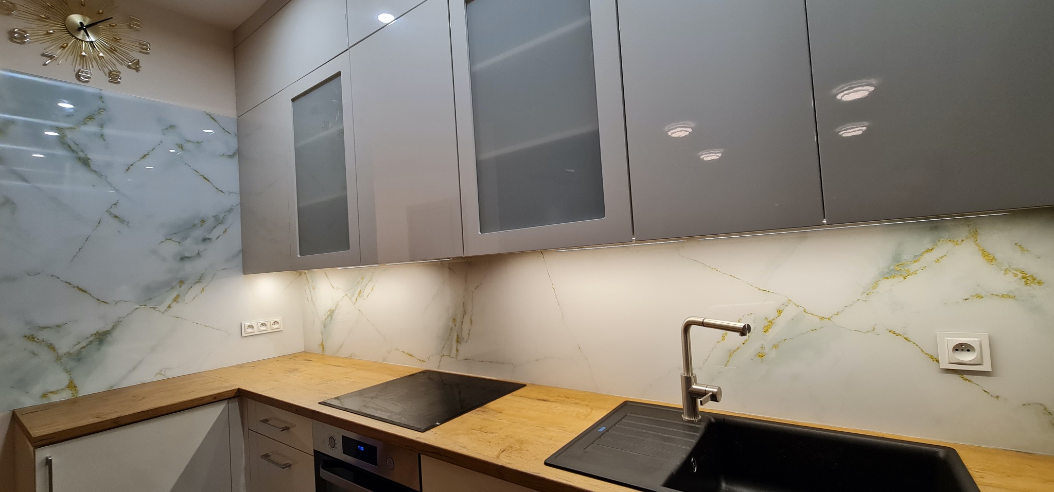 Nowoczesna kuchnia z szarymi szafkami, drewnianym blatem i marmurowym panelem ściennym z złotymi żyłkami. Oświetlenie LED pod szafkami górnymi.