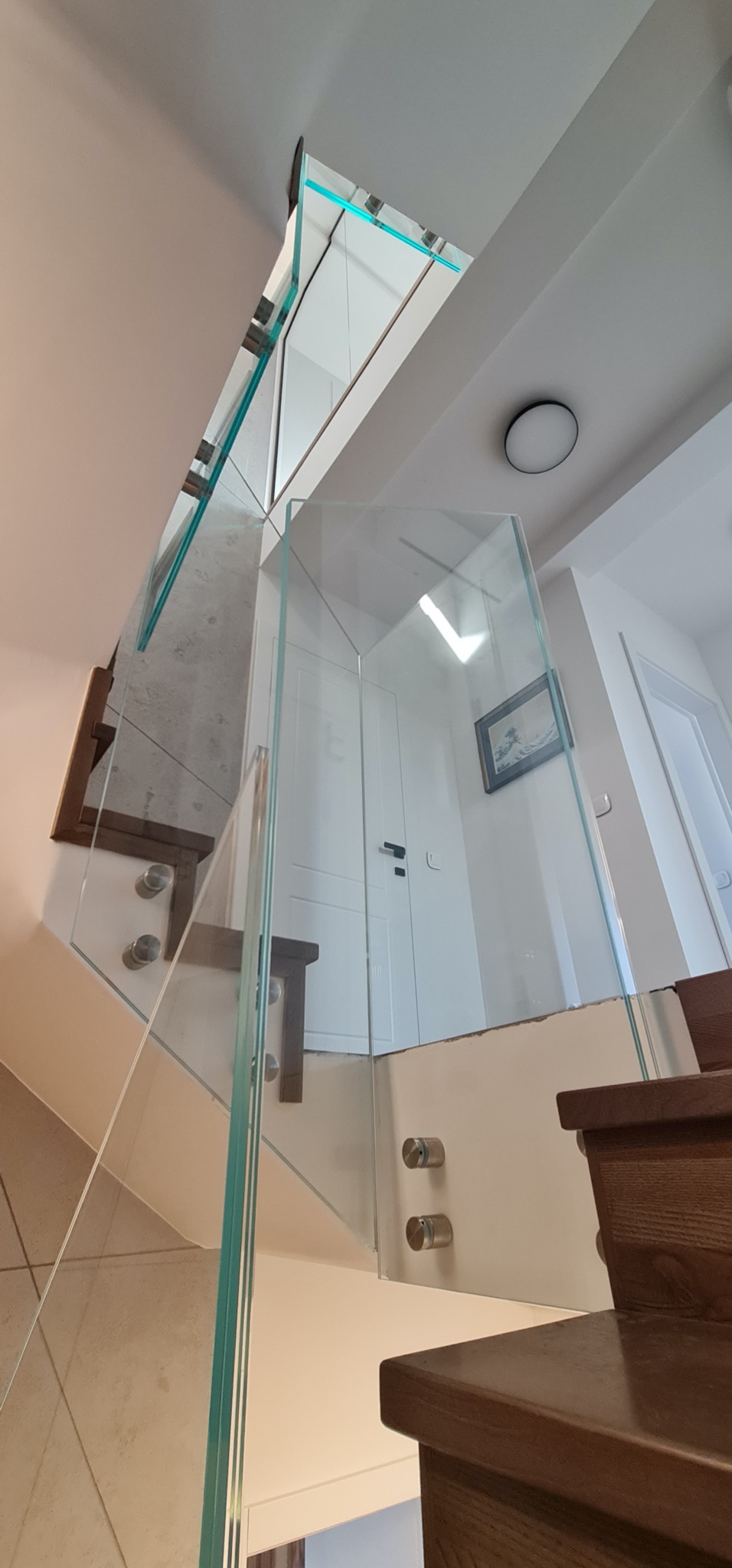 Szklana balustrada schodowa z drewnianymi stopniami, mocowana na metalowych uchwytach. Widok z dołu, ukazujący przejrzystość szkła i nowoczesny design.