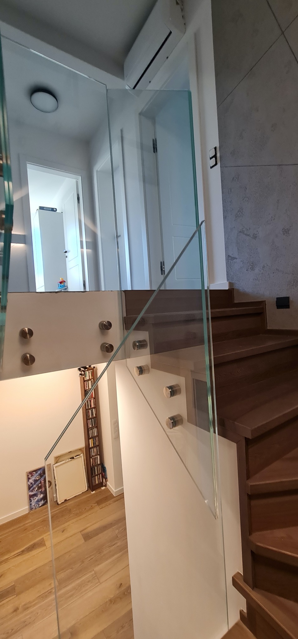 Szklana balustrada schodowa z drewnianymi stopniami, mocowana na metalowych uchwytach. Widok na korytarz z białymi drzwiami i podłogą z paneli. Nowoczesne wykończenie wnętrza.