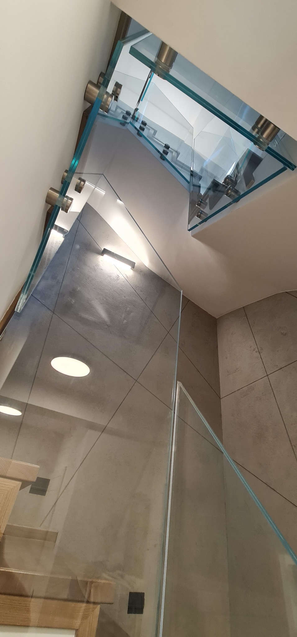 Szklana balustrada schodowa z metalowymi mocowaniami, widok z dołu, nowoczesny design, szare płytki na ścianie, oświetlenie punktowe.