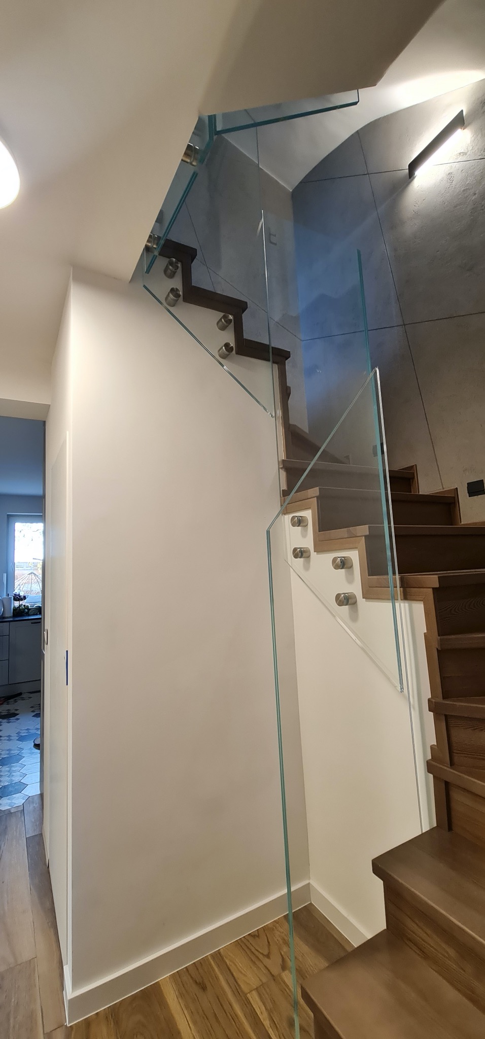 Nowoczesne schody z drewnianymi stopniami i szklaną balustradą mocowaną na metalowych uchwytach. Widok z dołu, jasne wnętrze.