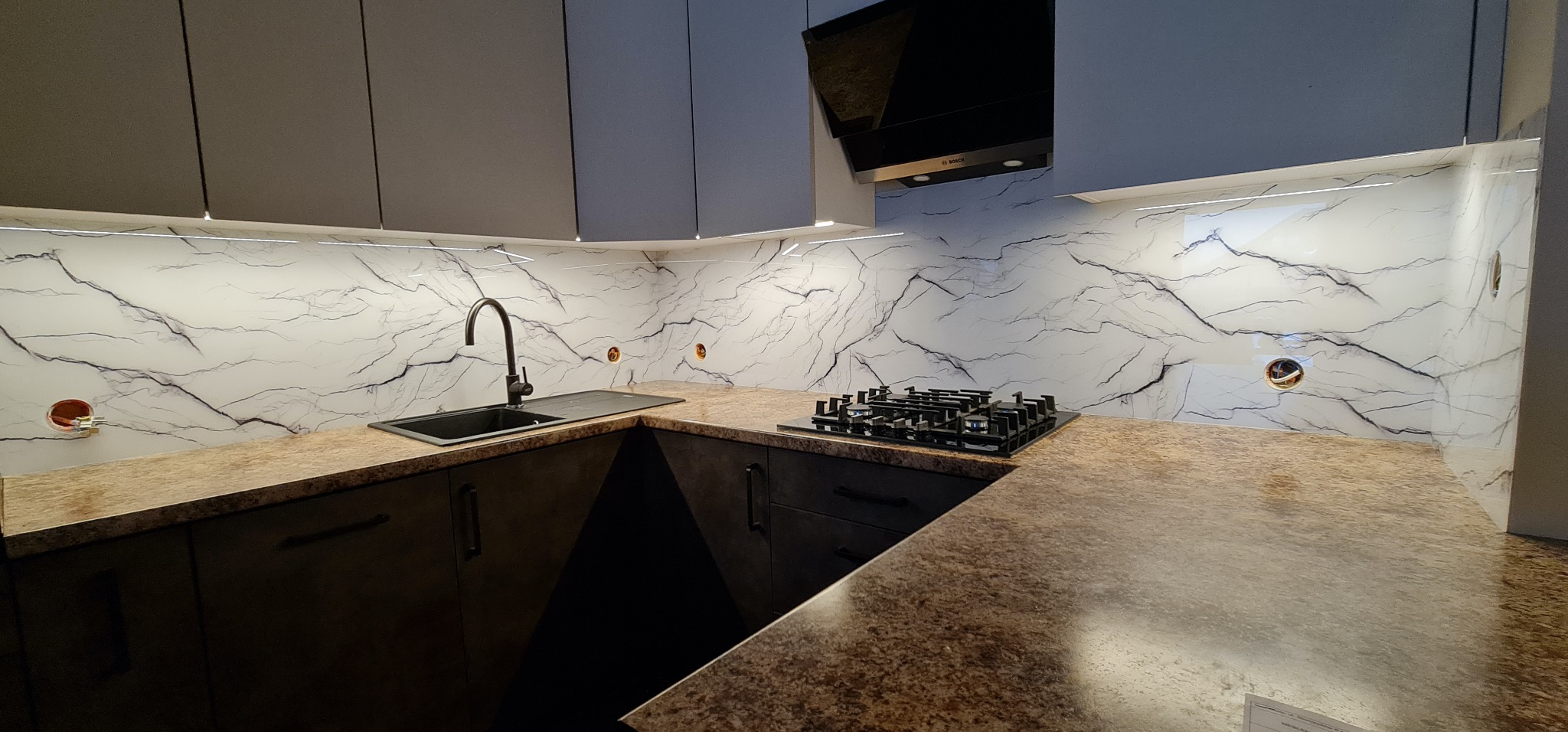 Nowoczesna kuchnia z marmurowym panelem szklanym nad blatem, oświetleniem LED i czarną armaturą. Widoczne gniazdka elektryczne i płyta gazowa.