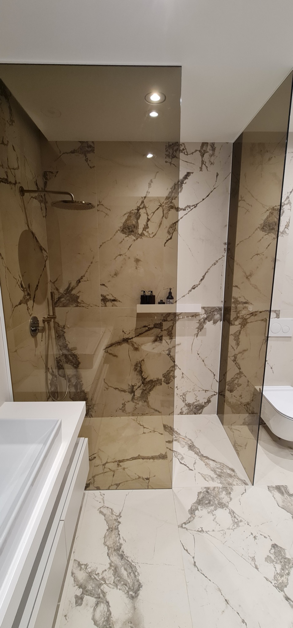 Nowoczesna łazienka z kabiną typu walk-in, wykończona płytkami imitującymi marmur. Widoczny zestaw prysznicowy i umywalka z szafką.