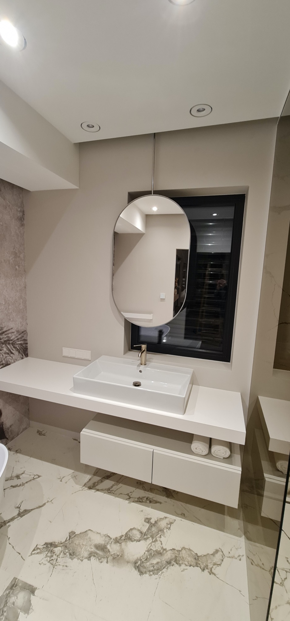 Nowoczesna łazienka z owalnym lustrem wiszącym na metalowym pręcie, białą umywalką nablatową i marmurową podłogą. Minimalistyczny design, ciepłe oświetlenie.