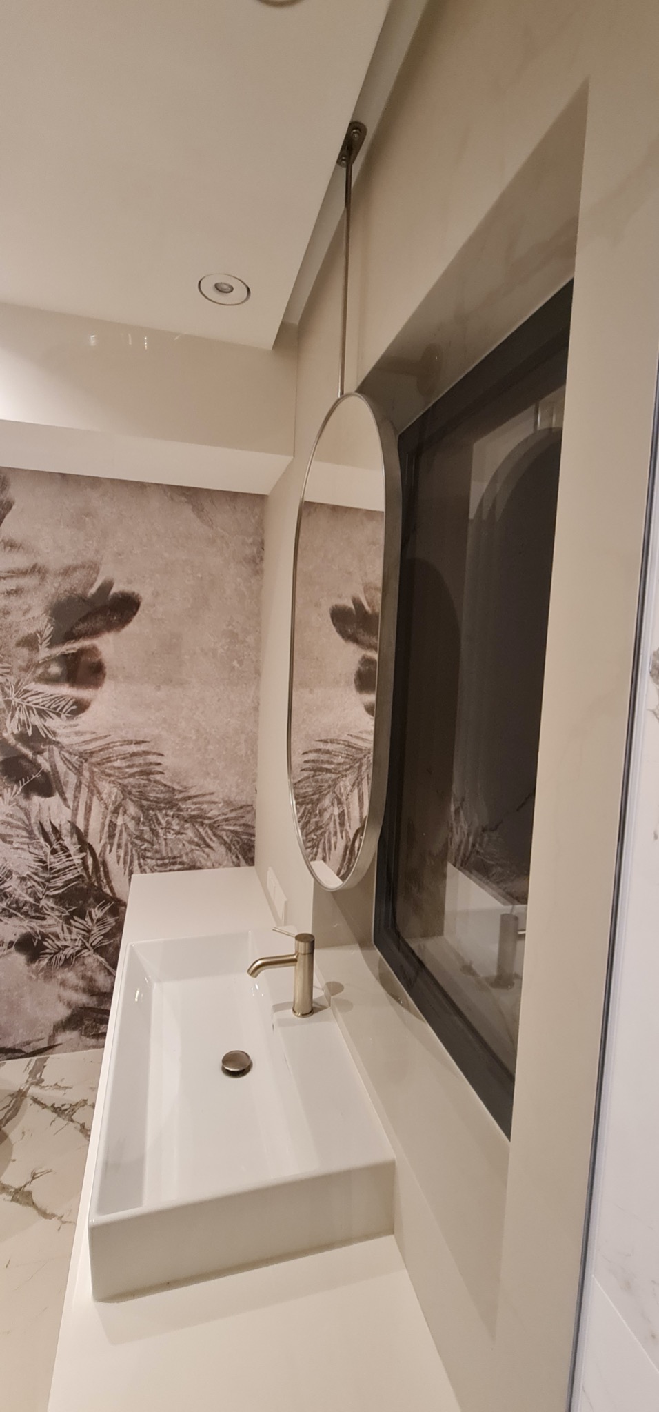 Nowoczesna łazienka z owalnym lustrem na metalowym stelażu, białą umywalką i baterią w kolorze szczotkowanego złota. Na ścianie tapeta z motywem roślinnym.