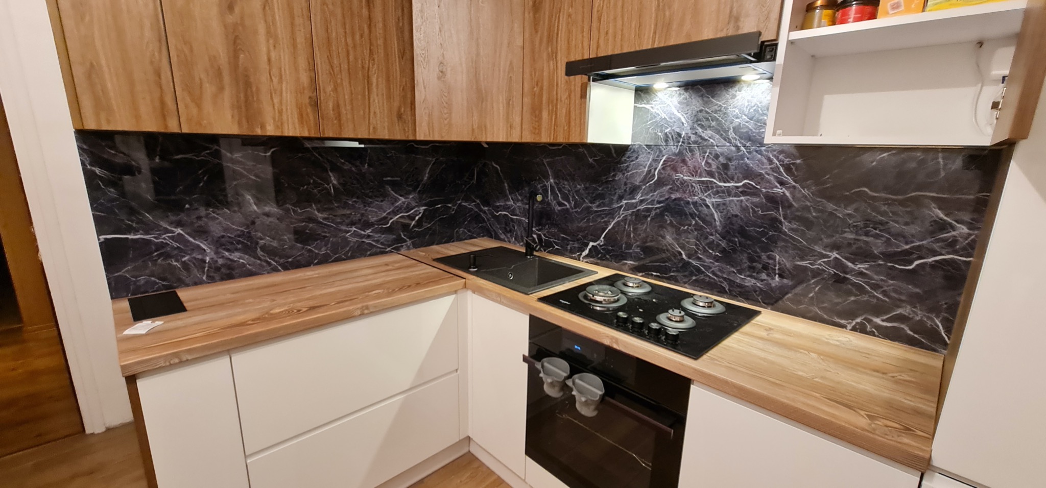 Nowoczesna kuchnia z białymi szafkami, drewnianym blatem i czarnym panelem ściennym imitującym marmur. Widoczny zlew, płyta gazowa i piekarnik.