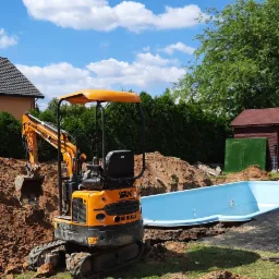 Pomarańczowa mini-koparka Berger Kraus BK1000AS w trakcie prac ziemnych przy wykopie pod basen z niebieską wkładką, na trawniku, z widocznym niebem i chmurami.