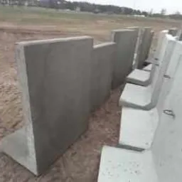 Szereg prefabrykowanych, betonowych elementów w kształcie litery L, ustawionych na ziemi. Widoczne metalowe uchwyty w niektórych elementach.