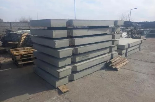 Stos szarych, betonowych płyt drogowych o prostokątnym przekroju, ułożonych na placu budowy w pochmurny dzień, z widocznymi paletami i innymi materiałami budowlanymi w tle.