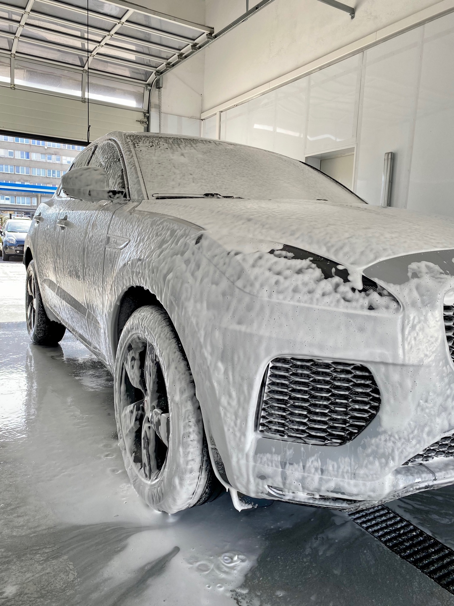 Szary SUV pokryty gęstą pianą w myjni samochodowej, widok z przodu na koło i kratkę wlotu powietrza, otwarte drzwi garażowe w tle.
