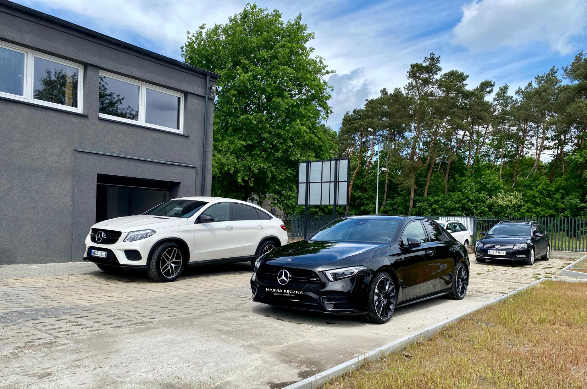 Dwa samochody marki Mercedes, biały SUV i czarny hatchback, zaparkowane przed budynkiem o szarej elewacji; w tle widoczny fragment parkingu i drzewa.
