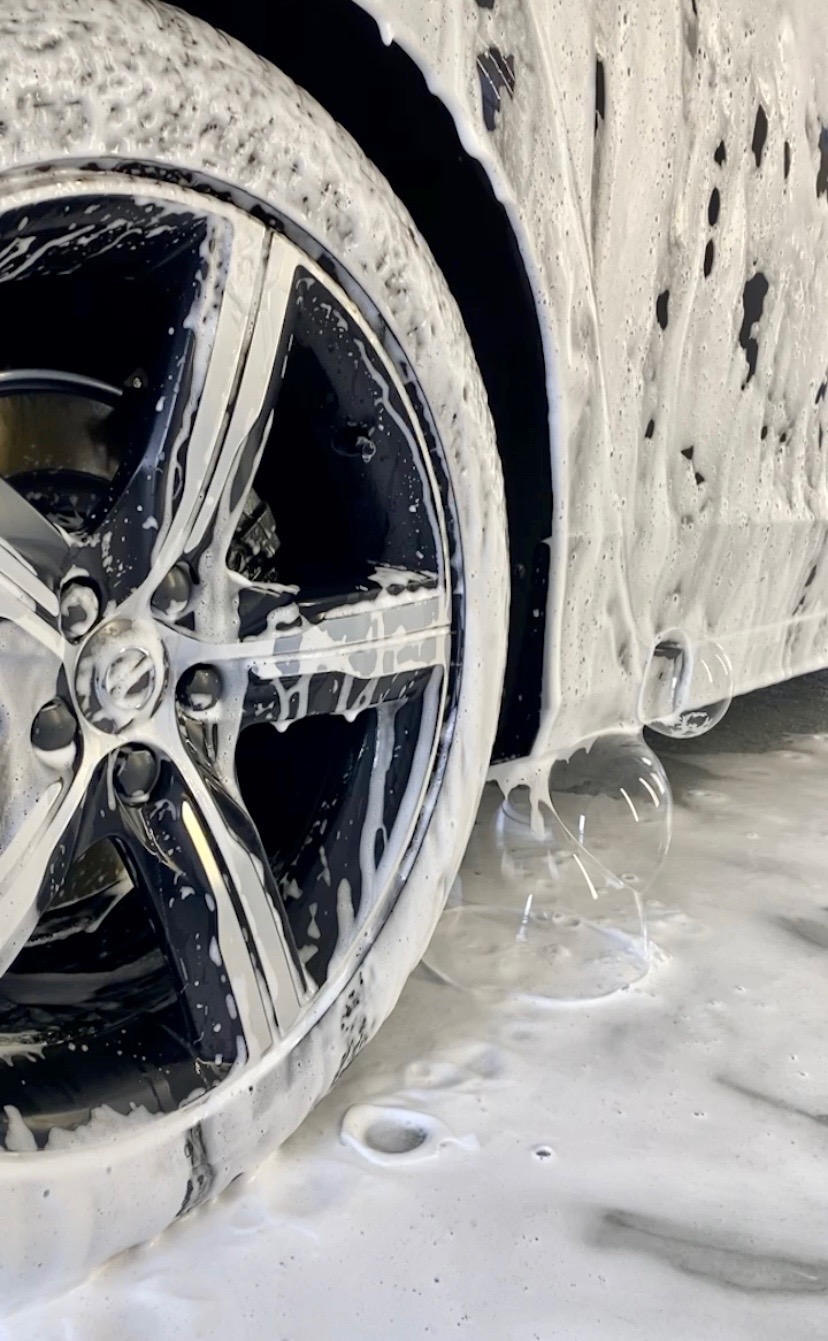 Czyszczone felgi aluminiowe samochodu pokryte grubą warstwą piany aktywnej, widoczne krople spływającej wody.
