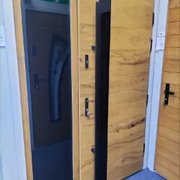 Wystawa drzwi z drewnianym fornirem i czarnym, pionowym panelem na skrzydle, z widocznym zamkiem i kluczem, obok drzwi z przeszkleniem, prezentowane w salonie.