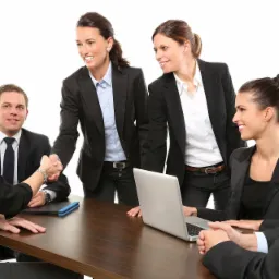 Grupa uśmiechniętych profesjonalistów w garniturach siedzi przy stole konferencyjnym podczas spotkania biznesowego, gdzie jedna osoba wita się uściskiem dłoni z inną, a na stole leży otwarty laptop.