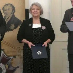 Kobieta w czarnej marynarce i sukience w kropki, z odznaczeniami, trzymająca niebieski folder z symbolem gwiazdy na tle portretu.