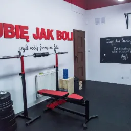 Wnętrze siłowni z czarną podłogą, czerwonymi akcentami, sprzętem do ćwiczeń, ścianą z napisem 'Lubię jak boli czyli robię formę na lata' i tablicą z planem treningowym oraz ofertą batonów...
