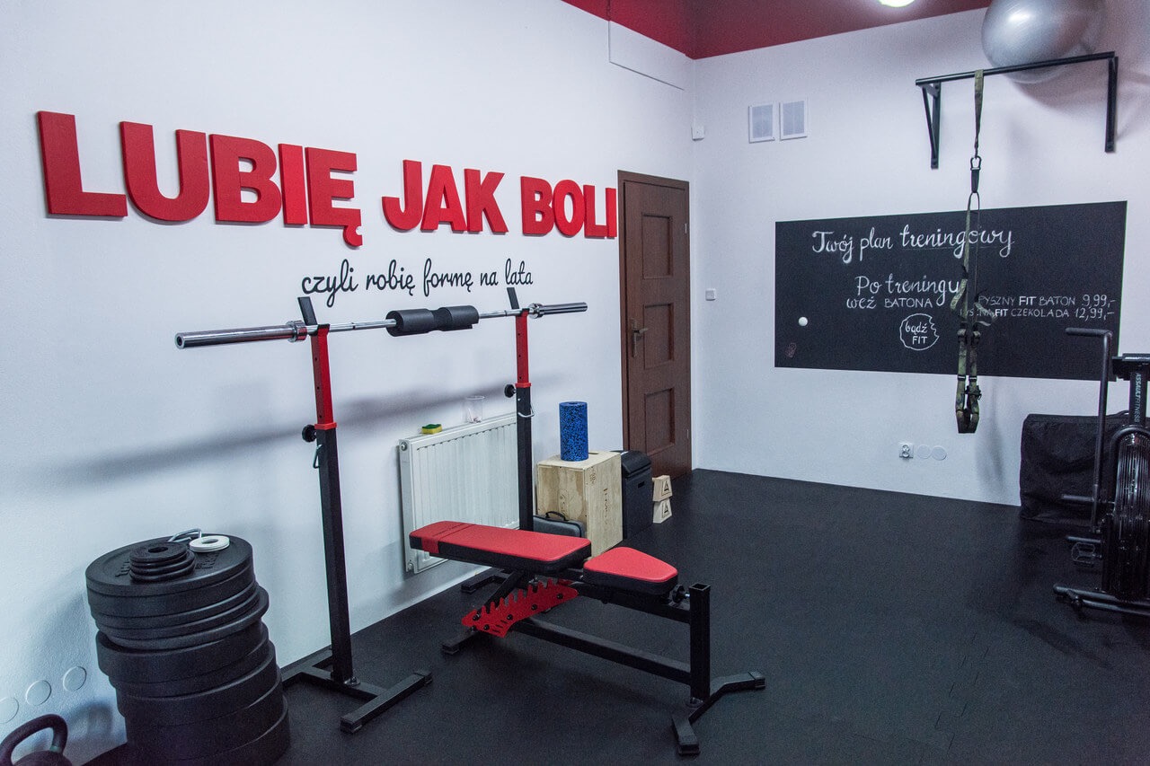 Wnętrze siłowni z czarną podłogą, czerwonymi akcentami, sprzętem do ćwiczeń, ścianą z napisem 'Lubię jak boli czyli robię formę na lata' i tablicą z planem treningowym oraz ofertą batonów...