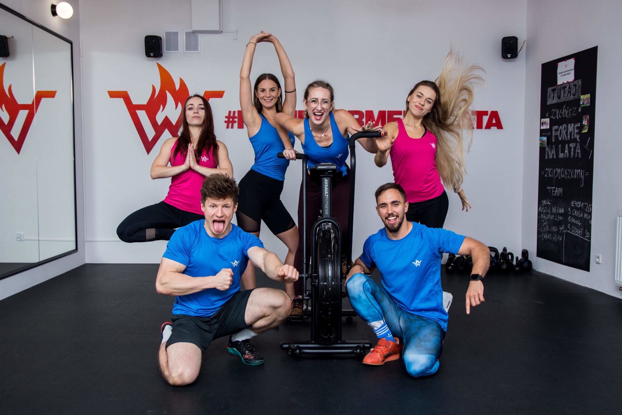 Grupa uśmiechniętych osób w sportowych strojach pozuje w siłowni w Krakowie, z rowerem treningowym na pierwszym planie i tablicą z hasłami motywacyjnymi w tle.