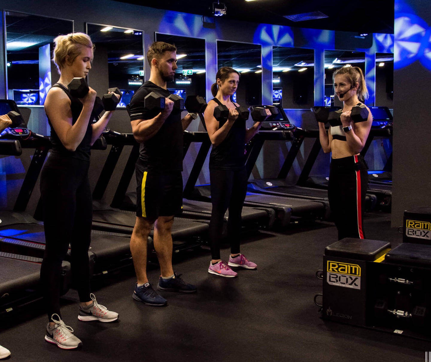 Trenerka personalna z mikrofonem instruuje grupę osób ćwiczących z hantlami w siłowni z bieżniami w tle i boksami RAM.