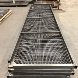 Stos metalowych krat pomostowych z kwadratowej rury, spawanych w narożach, ułożonych na betonowej podłodze w hali produkcyjnej.