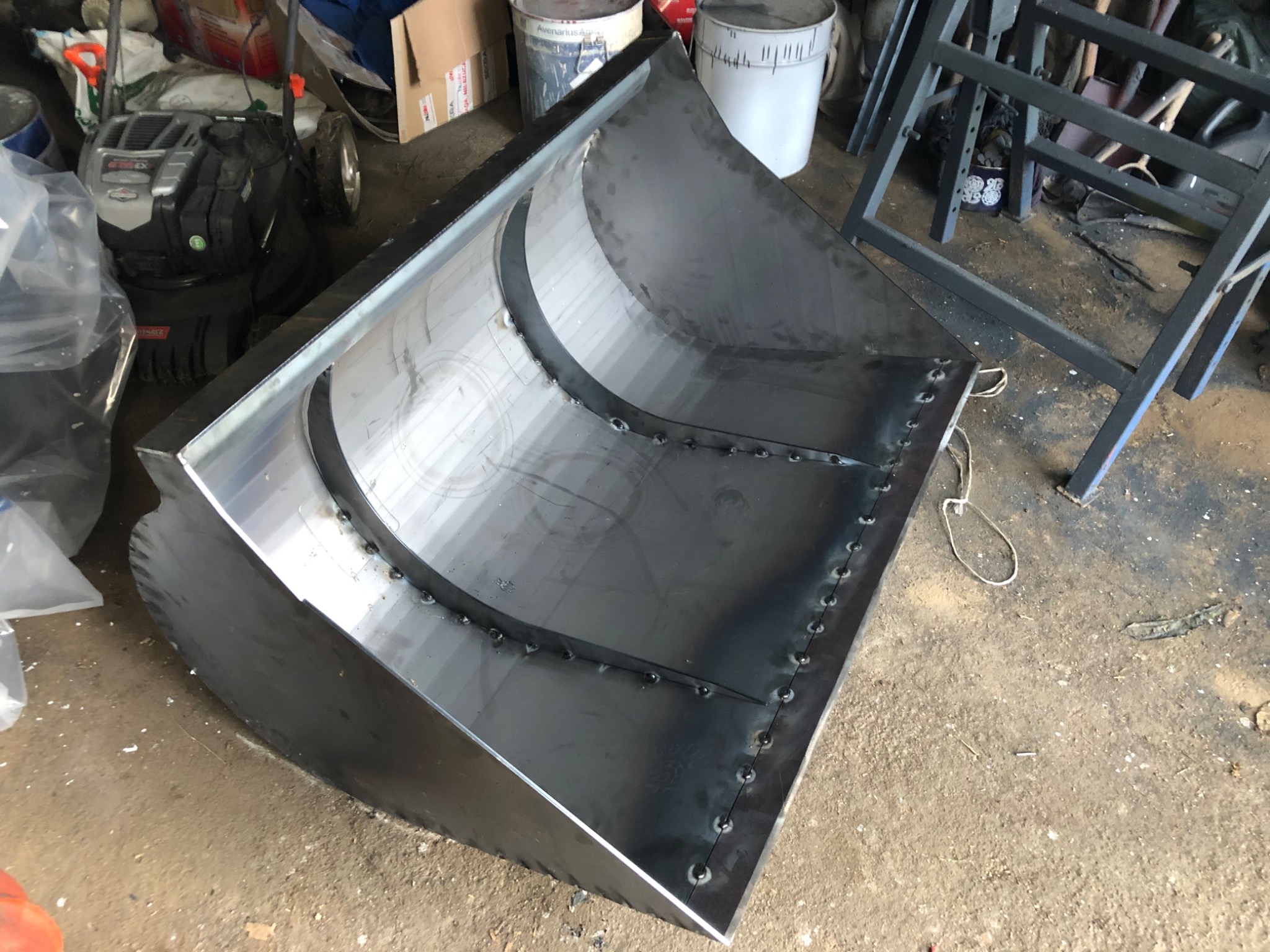 Metalowa konstrukcja przypominająca łyżkę koparki, spawana punktowo, widoczna w warsztacie, obok kosiarka i inne sprzęty.