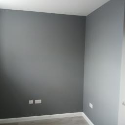 Pomalowane na szaro wnętrze pokoju, widoczne gładkie ściany, białe gniazdka elektryczne i fragment podłogi.