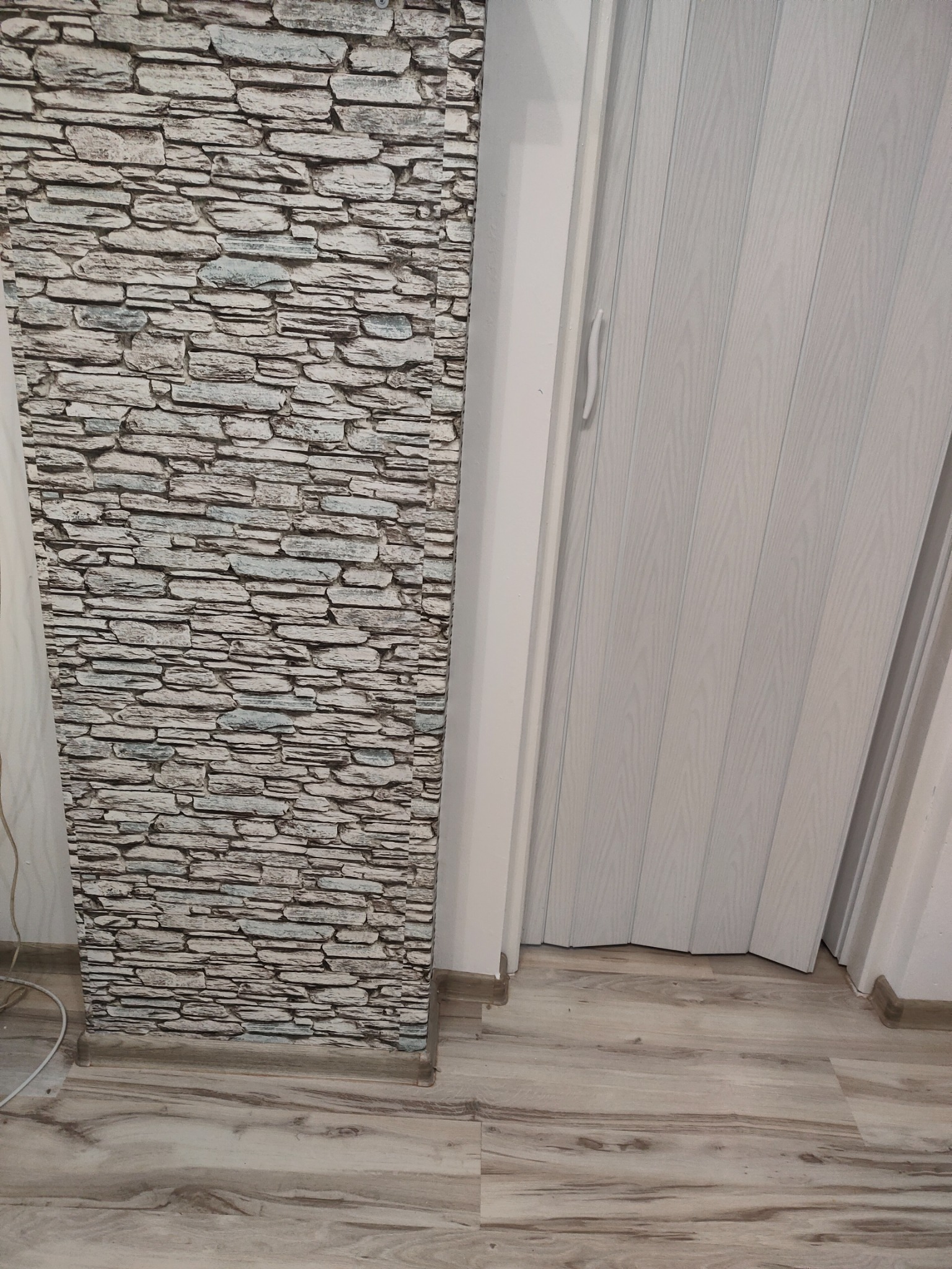 Fragment ściany z tapetą imitującą kamień, przylegający do białych, składanych drzwi harmonijkowych na podłodze z paneli imitujących drewno.