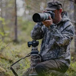 Fotograf przyrody w kamuflażu, klęczący w lesie z aparatem na statywie i drugim aparatem w rękach, robiący zdjęcie.