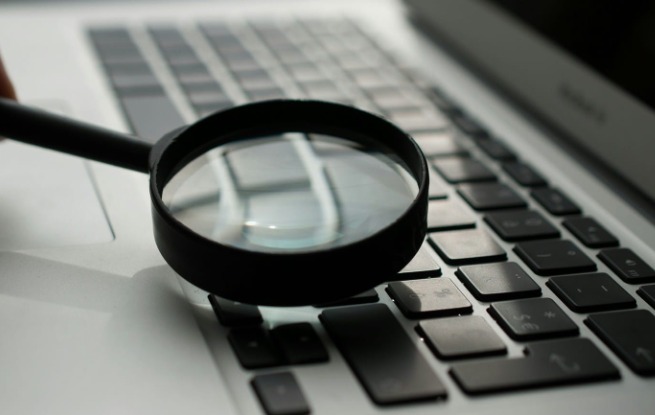 Czarna lupa powiększa klawiaturę laptopa, delikatnie rozmyte tło, sugerujące poszukiwanie informacji lub analizę danych.