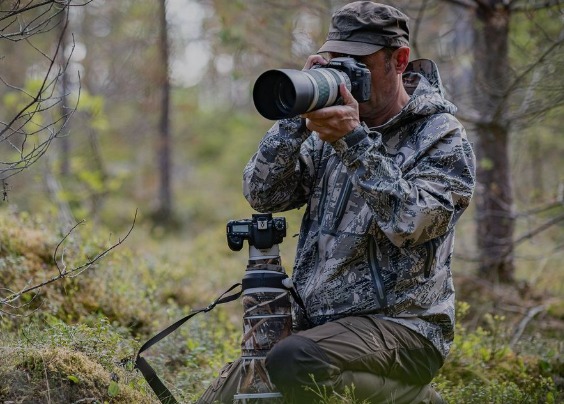 Fotograf przyrody w kamuflażu, klęczący w lesie z aparatem na statywie i drugim aparatem w rękach, robiący zdjęcie.
