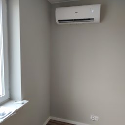 Vector Serwis - Biały klimatyzator Haier zamontowany na szarej ścianie w pokoju z drewnianą podłogą, widoczny wyświetlacz temperatury 13 stopni, obok okno z pilotem na parapecie.