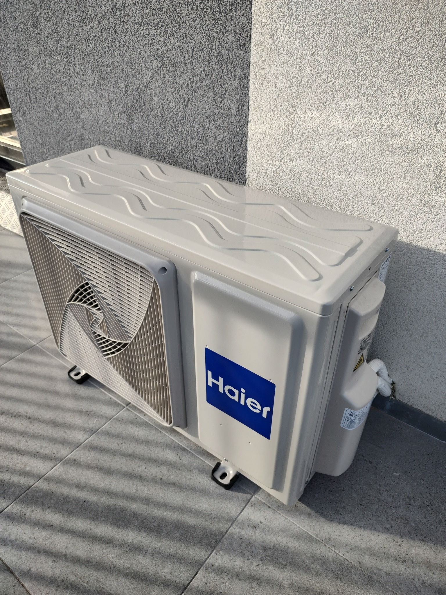 Zewnętrzna jednostka klimatyzacji Haier zamontowana na szarym tarasie, widoczna na tle ściany o fakturze baranka. Na obudowie niebieskie logo producenta.