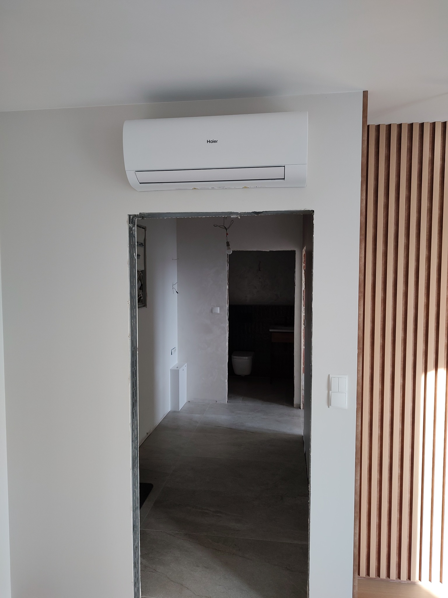 Biały klimatyzator Haier zamontowany nad otworem drzwiowym w trakcie remontu, widoczne drewniane lamele dekoracyjne na ścianie i wnętrze pomieszczenia w głębi.
