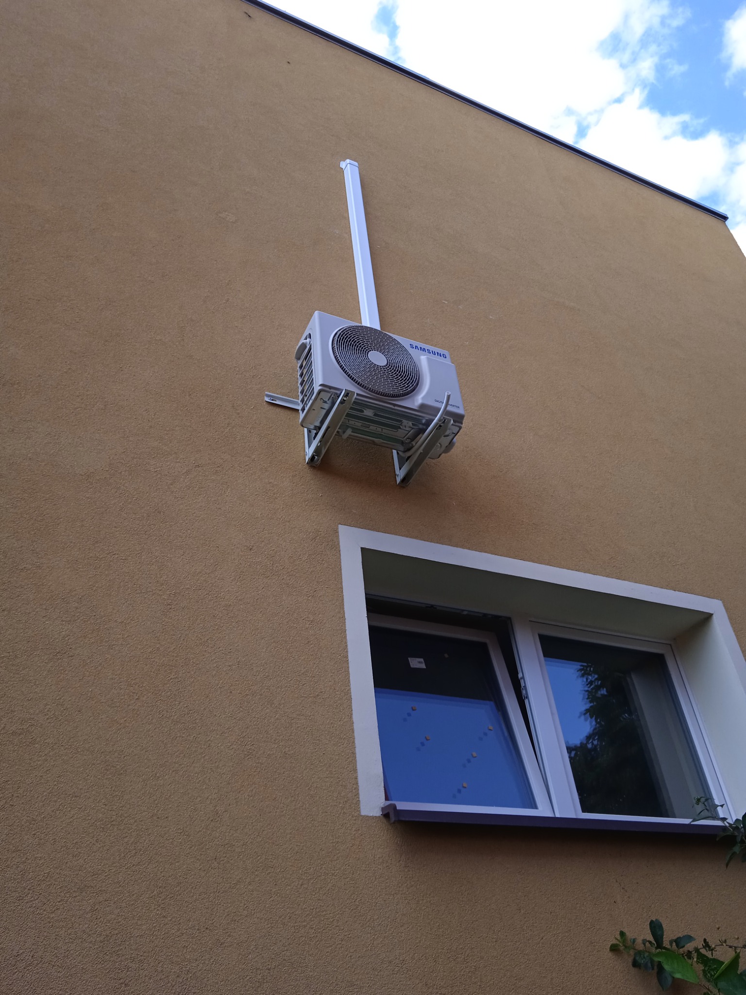Zewnętrzna jednostka klimatyzacji Samsung zamontowana na elewacji budynku obok okna, z widocznymi elementami montażowymi i rurą odprowadzającą.
