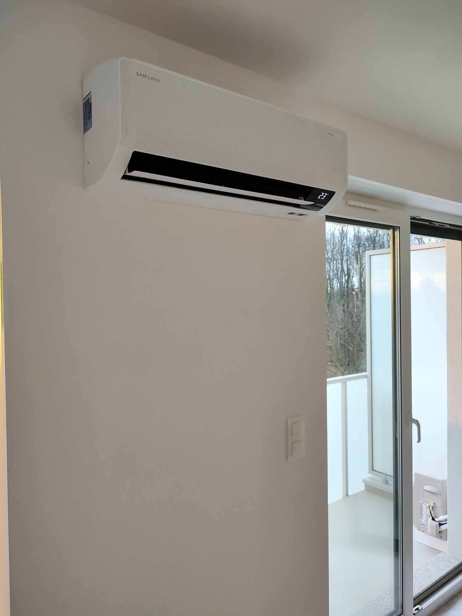 Biała jednostka wewnętrzna klimatyzacji Samsung zamontowana na ścianie w jasnym pomieszczeniu, widoczny balkon zza przesuwanych drzwi balkonowych.