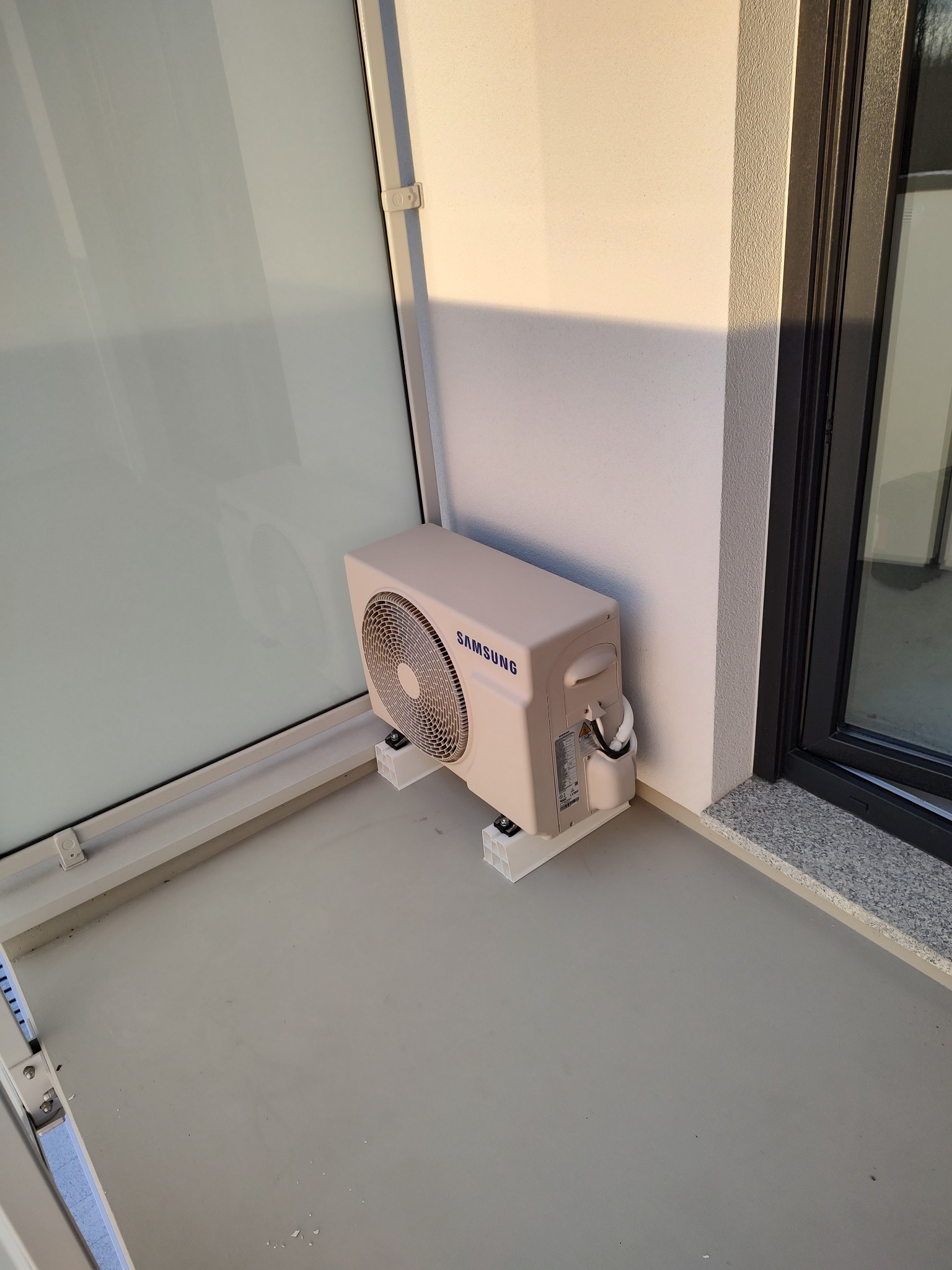 Zewnętrzna jednostka klimatyzacji Samsung zamontowana na wspornikach z tworzywa sztucznego na balkonie, widok z góry.