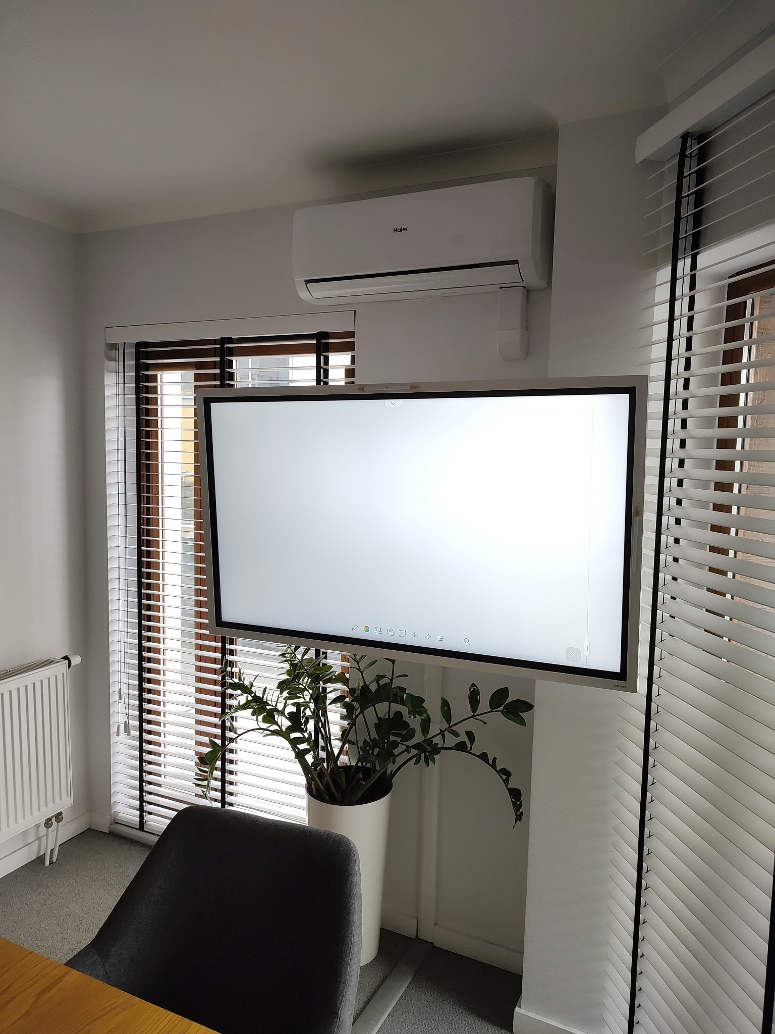 Biała klimatyzacja Haier zamontowana nad interaktywną tablicą w jasnym biurze z żaluzjami, obok doniczka z zieloną rośliną.