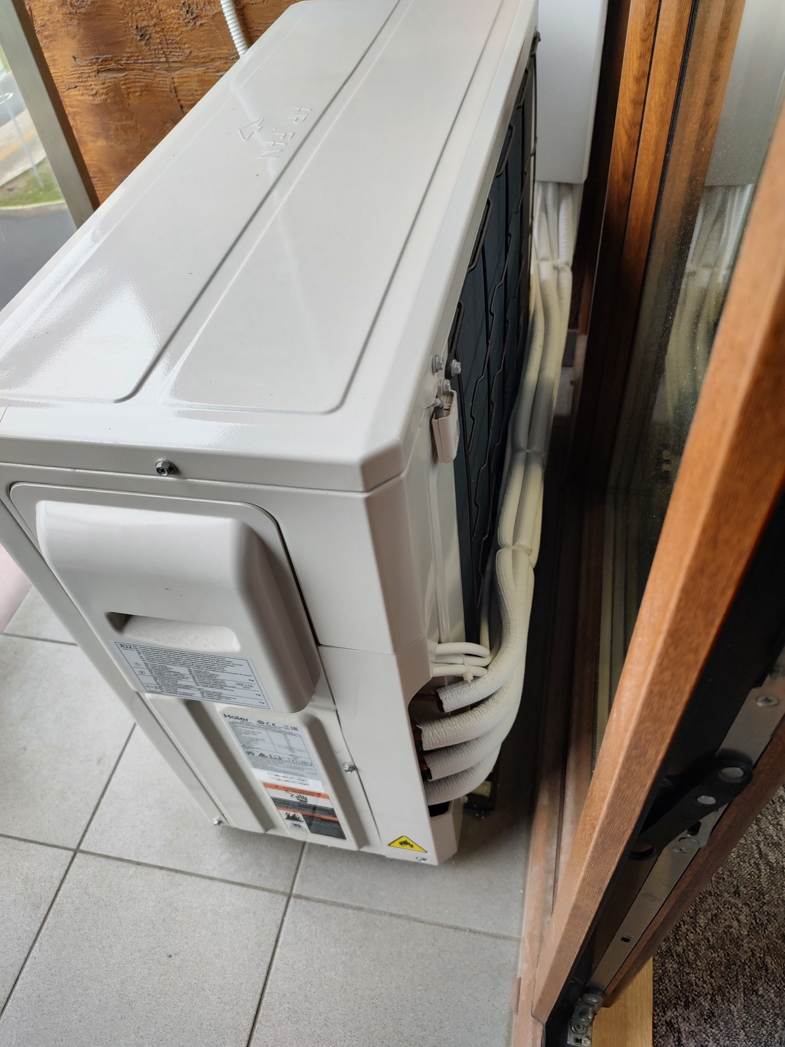 Boczny widok białej jednostki zewnętrznej klimatyzacji Haier, zamontowanej na balkonie, z widocznymi przewodami i kratką wentylacyjną, oraz fragmentem drewnianego okna.