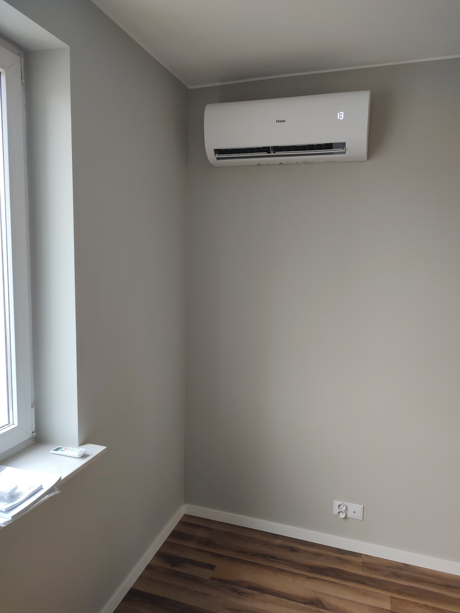 Biały klimatyzator Haier zamontowany na szarej ścianie w pokoju z drewnianą podłogą, widoczny wyświetlacz temperatury 13 stopni, obok okno z pilotem na parapecie.