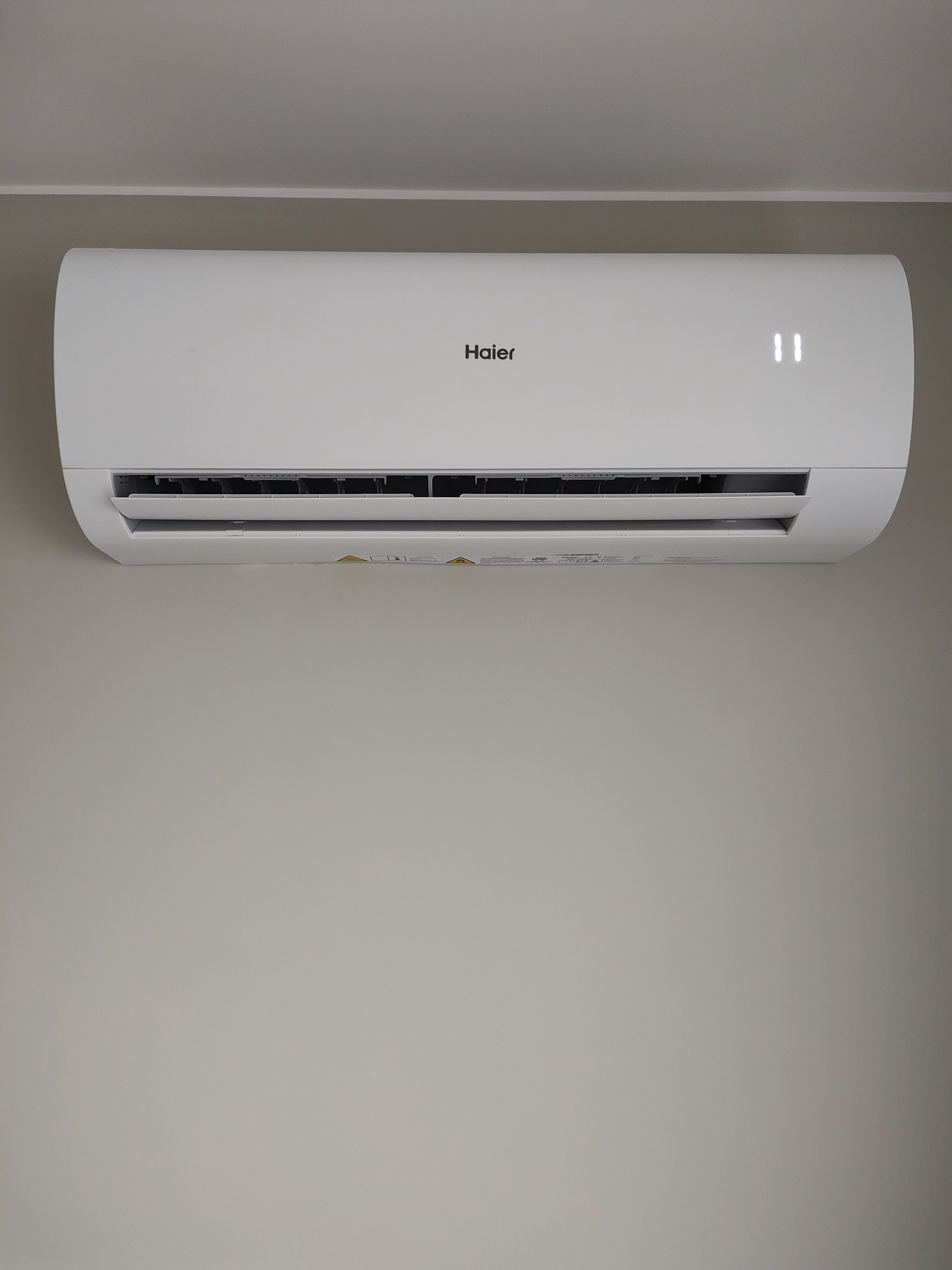 Biała klimatyzacja Haier zamontowana na szarej ścianie, wyświetlająca temperaturę 11 stopni.