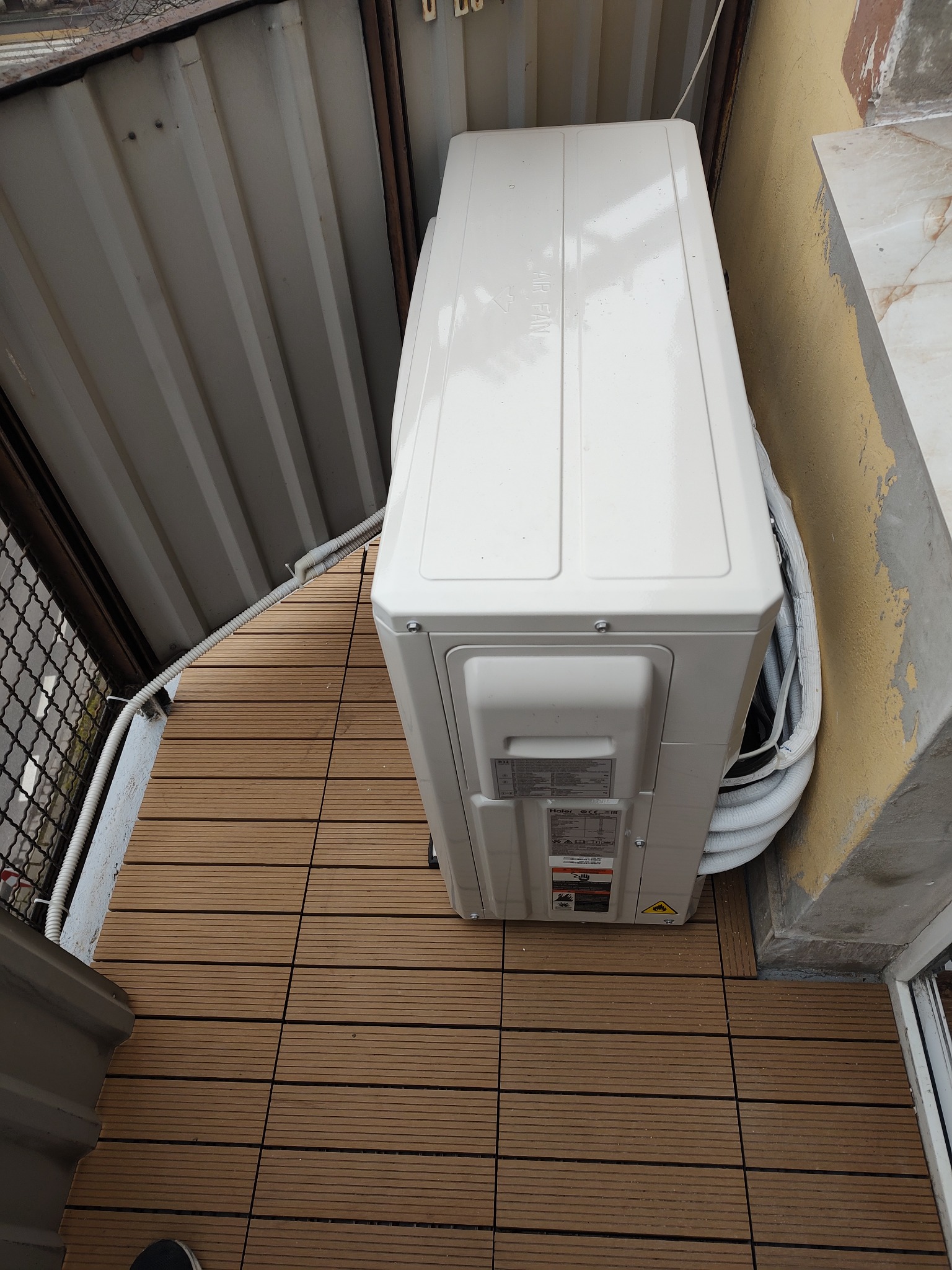 Zewnętrzna jednostka klimatyzacji Haier zamontowana na balkonie z brązową podłogą kompozytową, widoczne rury w izolacji termicznej i fragment ściany budynku.