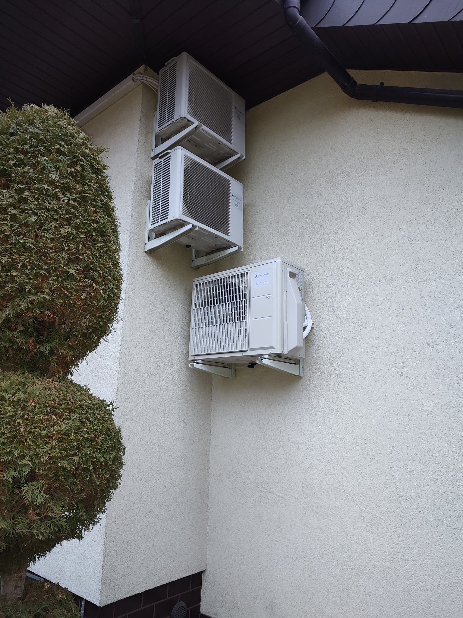 Trzy jednostki klimatyzacji zamontowane jedna nad drugą na elewacji budynku, obok ozdobnego krzewu.