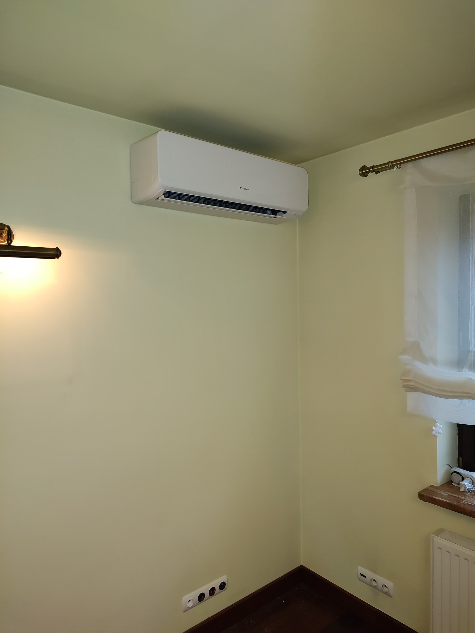 Biały klimatyzator ścienny zamontowany w rogu pokoju o jasnożółtych ścianach, widoczne gniazdka elektryczne i fragment grzejnika pod oknem.