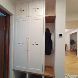 Biała szafa w przedpokoju z dekoracyjnymi wycięciami w kształcie krzyżyków na frontach, złotymi uchwytami i otwartą wnęką z wieszakiem na ubrania. Pod szafą półki na buty, a na podłodze wzorzysta...